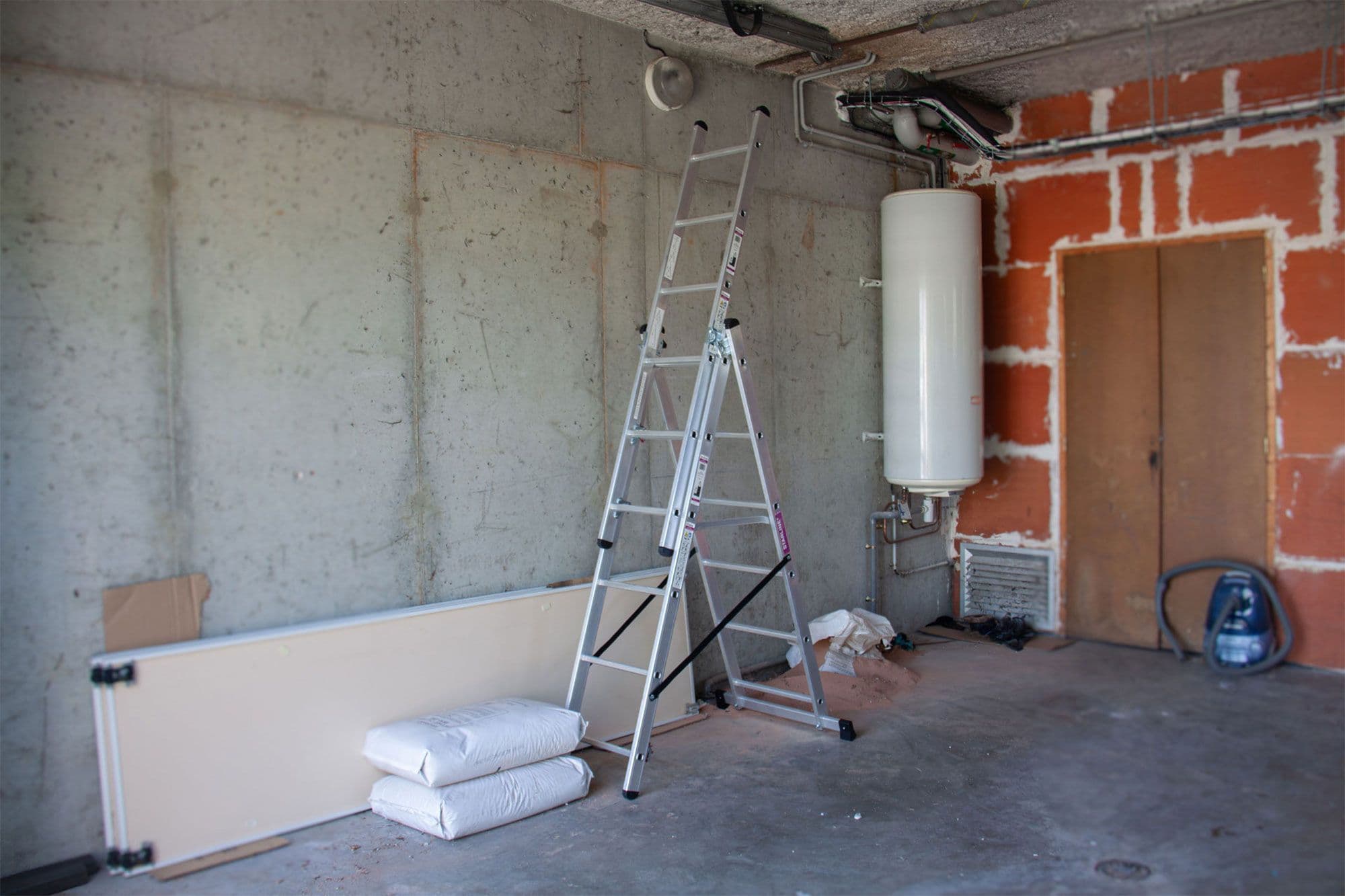 Unfinished room with a ladder, water heater, drywall sheet, and cement bags.