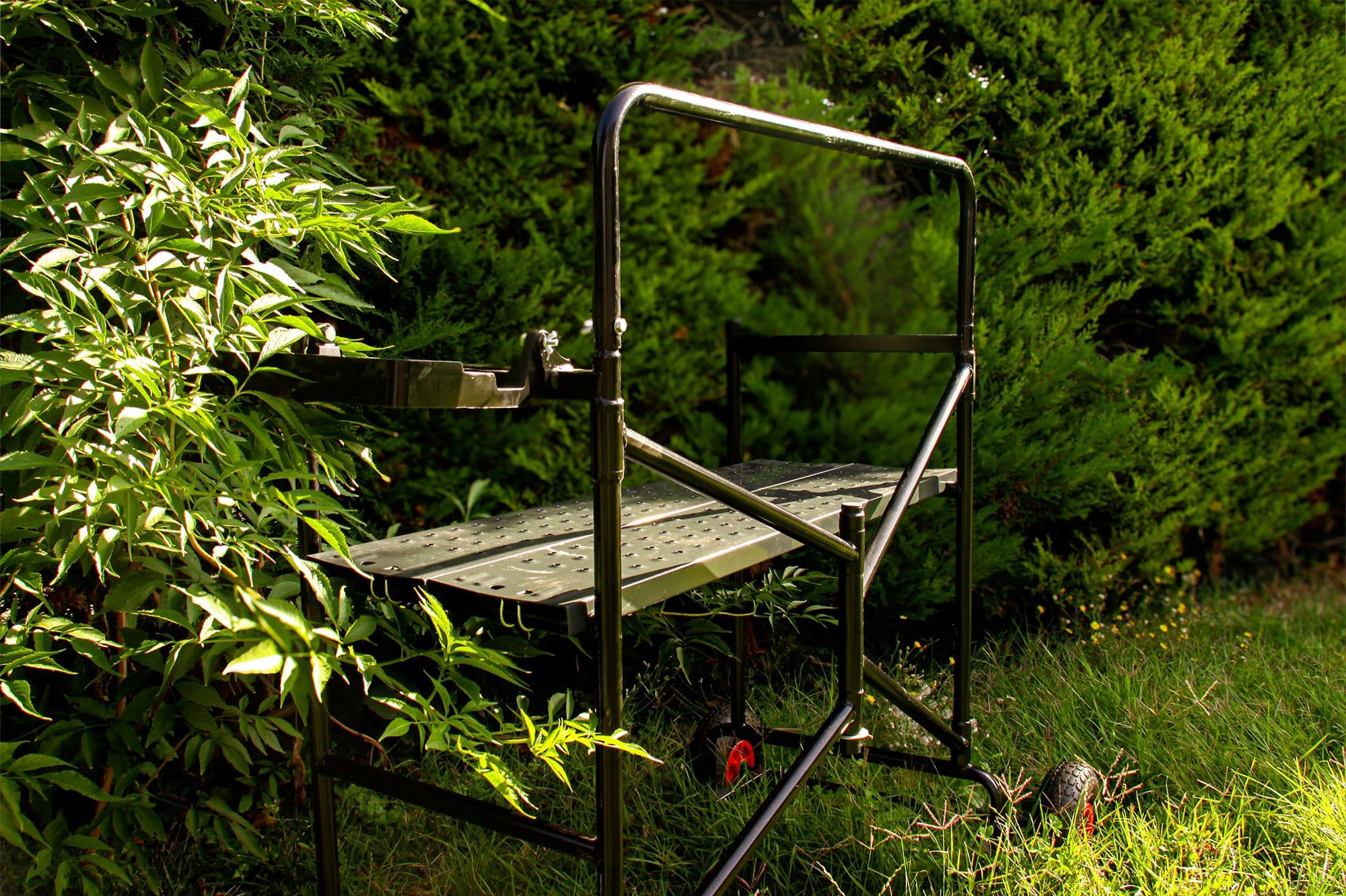 A portable black metal work platform stands in a sunlit garden among green foliage.